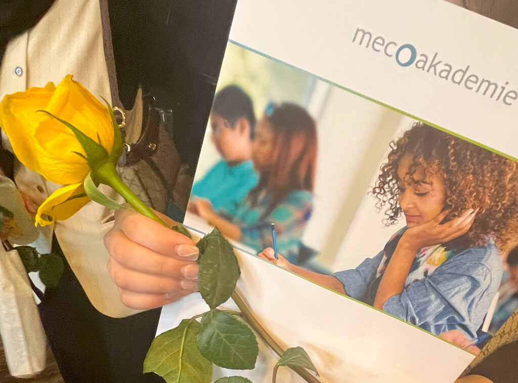 Zu sehen sind eine Mappe der meco Akademie, die zusammen mit einer Rose in den Händen gehalten wird.