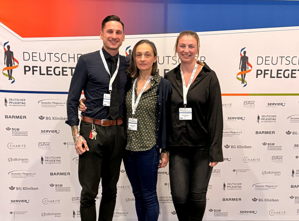Drei Mitarbeiter der meco Akademie stehen vor einem Banner des Deutschen Pflegetages 2025